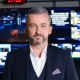 Krzysztof Skórzyński, elegancki mężczyzna w garniturze, stoi przed ścianą monitorów. Ile ma lat? Tego nie wiemy, ale wygląda na doświadczonego dziennikarza.