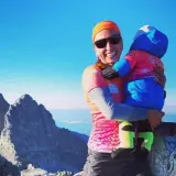 Justyna Kowalczyk z dzieckiem na szczycie górskim, uśmiechnięta, w słoneczny dzień.
