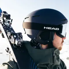 Mężczyzna w kasku narciarskim i goglach HEAD niesie narty. Różnice między kaskiem narciarskim a snowboardowym są subtelne, ale ważne dla bezpieczeństwa.