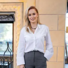 Uśmiechnięta Kamila Boś, której wiek zdaje się być nieistotny w jej profesjonalnym stroju, pozuje do zdjęcia w białej koszuli i szarych spodniach.