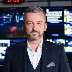 Krzysztof Skórzyński, elegancki mężczyzna w garniturze, stoi przed ścianą monitorów. Ile ma lat? Tego nie wiemy, ale wygląda na doświadczonego dziennikarza.