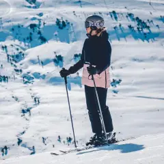 Narciarka w kasku i goglach stoi na stoku, trzymając kijki narciarskie.