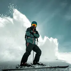 Narciarz w zielono-czarnej kurtce snowboardowej, z pomarańczowymi akcentami, w kasku i goglach, stoi na nartach, a za nim wybucha śnieżna lawina.