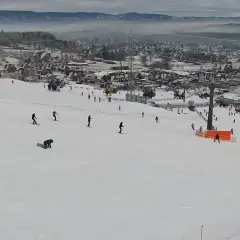 Widok z kamery na stok narciarski w Bodzentynie. Narciarze i snowboardziści szusują po ośnieżonych zboczach, w tle widoczne zabudowania i góry.