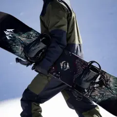 Osoba w zimowym stroju niesie dwa snowboardy z grafiką. Jeden z nich ma logo "JONES".