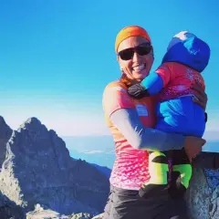 Justyna Kowalczyk z dzieckiem na szczycie górskim, uśmiechnięta, w słoneczny dzień.