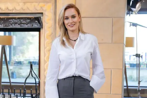 Uśmiechnięta Kamila Boś, której wiek zdaje się być nieistotny w jej profesjonalnym stroju, pozuje do zdjęcia w białej koszuli i szarych spodniach.