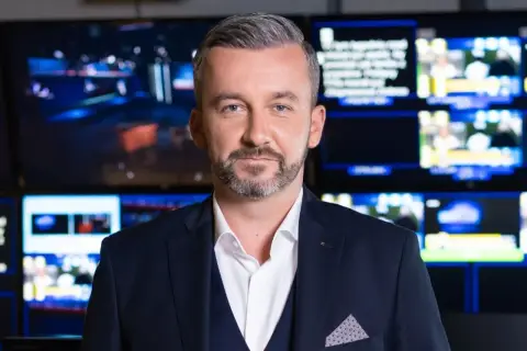 Krzysztof Skórzyński, elegancki mężczyzna w garniturze, stoi przed ścianą monitorów. Ile ma lat? Tego nie wiemy, ale wygląda na doświadczonego dziennikarza.