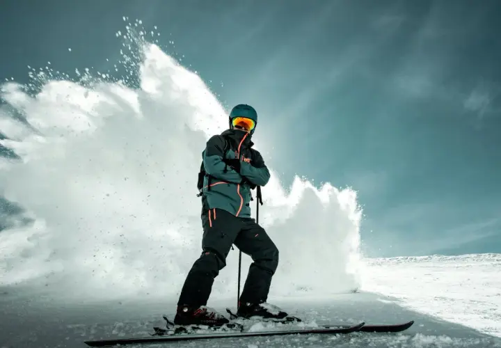 Narciarz w zielono-czarnej kurtce snowboardowej, z pomarańczowymi akcentami, w kasku i goglach, stoi na nartach, a za nim wybucha śnieżna lawina.