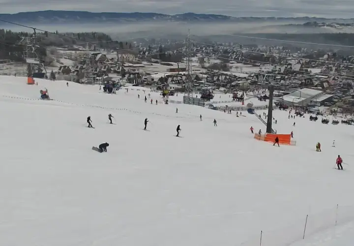 Widok z kamery na stok narciarski w Bodzentynie. Narciarze i snowboardziści szusują po ośnieżonych zboczach, w tle widoczne zabudowania i góry.