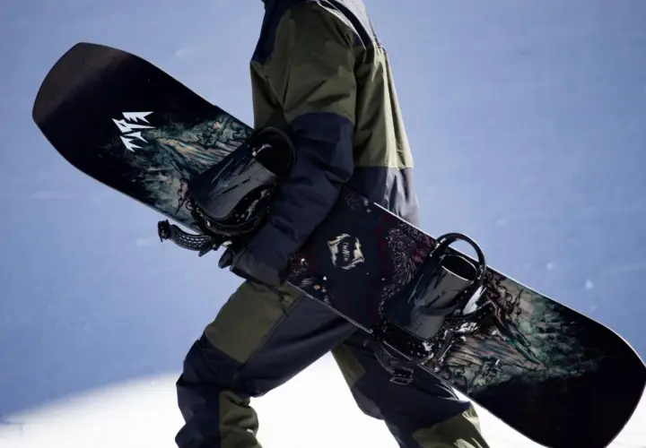 Osoba w zimowym stroju niesie dwa snowboardy z grafiką. Jeden z nich ma logo "JONES".