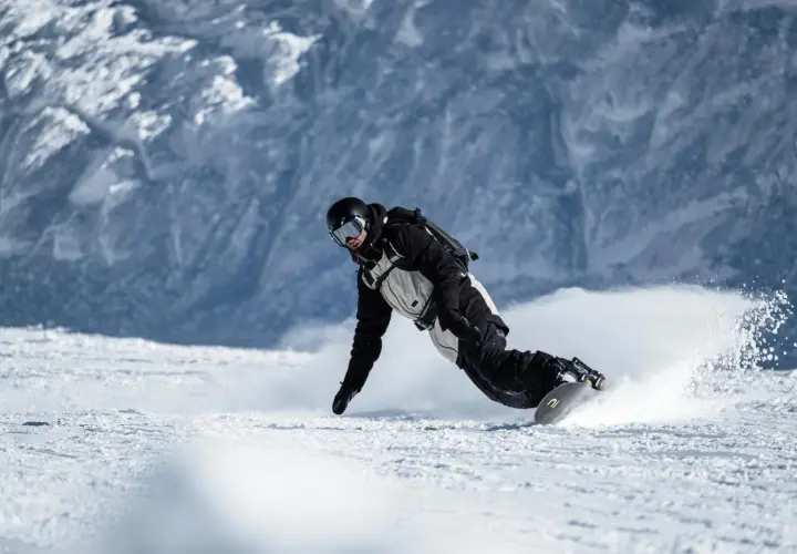 Snowboarder w kasku i goglach, z ochraniaczem nadgarstka snowboard, pochyla się nisko, tworząc obłok śniegu podczas zjazdu.
