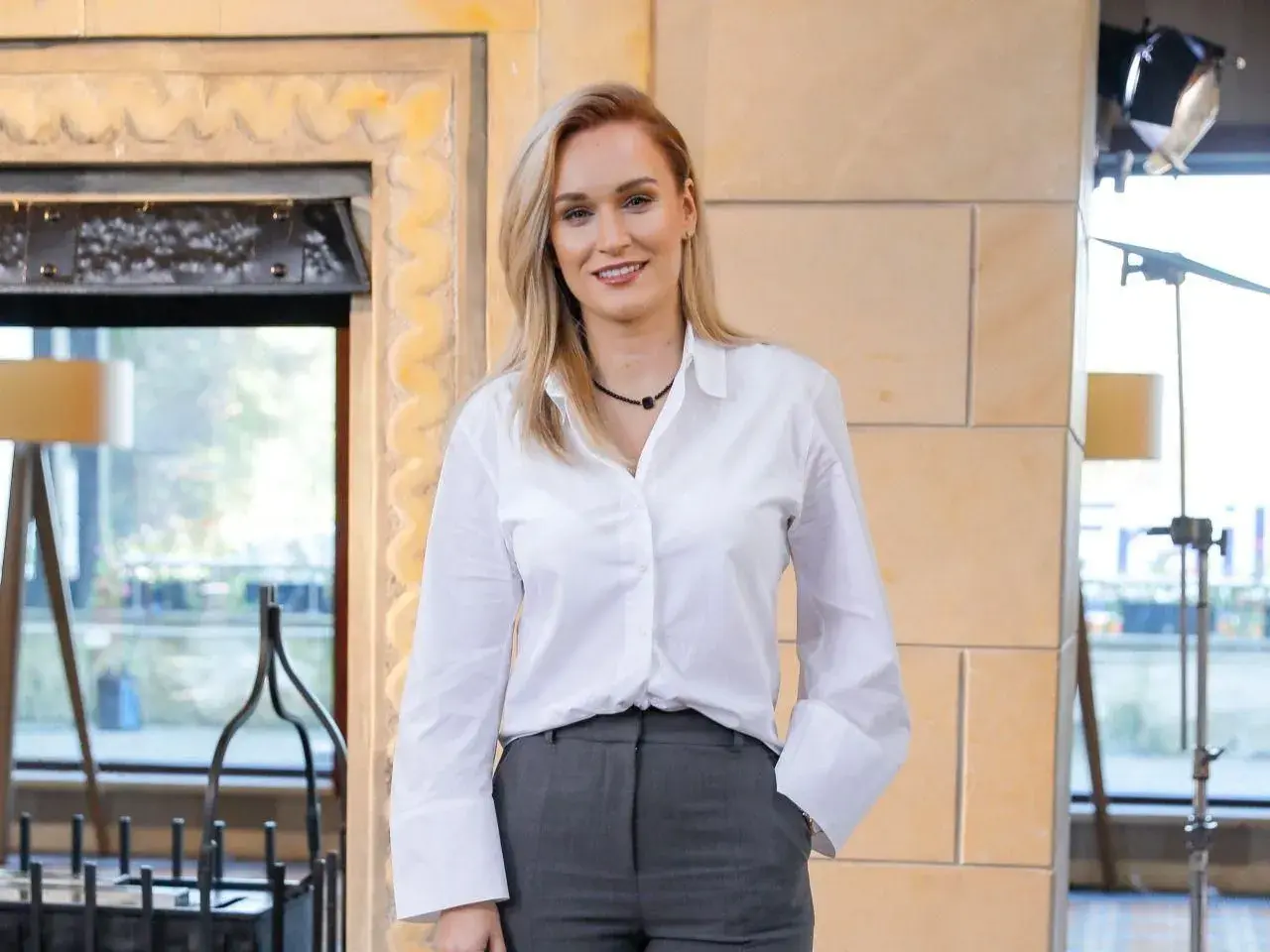 Uśmiechnięta Kamila Boś, której wiek zdaje się być nieistotny w jej profesjonalnym stroju, pozuje do zdjęcia w białej koszuli i szarych spodniach.