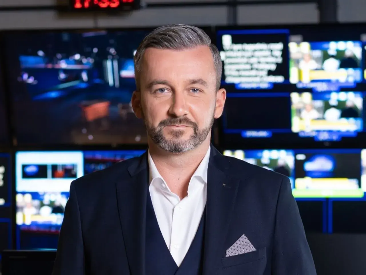 Krzysztof Skórzyński, elegancki mężczyzna w garniturze, stoi przed ścianą monitorów. Ile ma lat? Tego nie wiemy, ale wygląda na doświadczonego dziennikarza.