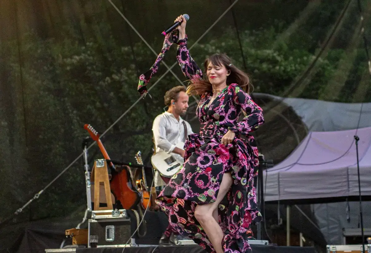 Bela Komoszyńska w kwiecistej sukni tańczy na scenie, trzymając mikrofon. W tle gitarzysta.