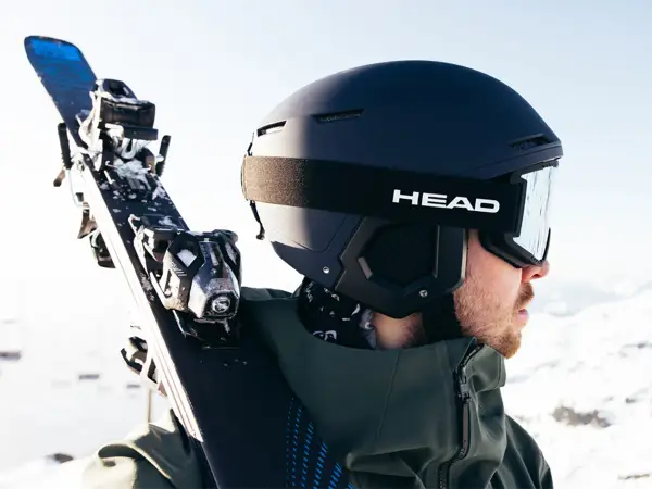 Mężczyzna w kasku narciarskim i goglach HEAD niesie narty. Różnice między kaskiem narciarskim a snowboardowym są subtelne, ale ważne dla bezpieczeństwa.
