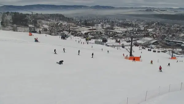 Widok z kamery na stok narciarski w Bodzentynie. Narciarze i snowboardziści szusują po ośnieżonych zboczach, w tle widoczne zabudowania i góry.